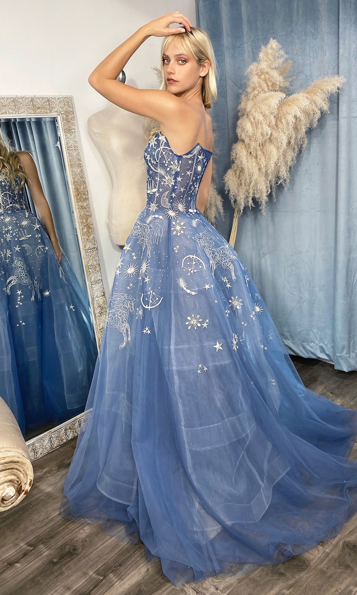 A woman wearing the Constellation-Print Strapless Prom Ball Gown A0890 stands before a mirror and a pampas grass arrangement.